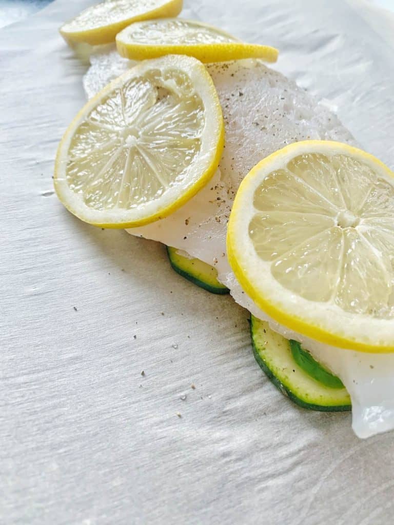 Slice of haddock on a piece of parchment paper with zucchini, lemon, salt and black pepper