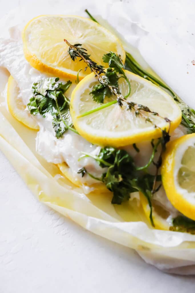 haddock fillet topped with sliced lemon, thyme and cilantro in a packet of parchment paper on a white backdrop