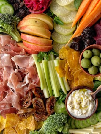close up shot of charcuterie board with prosciutto, chopped celery, sliced carrots, a bowl with coconut yogurt a wooden bowl with olives, sliced fights, chopped broccoli and sliced radishes