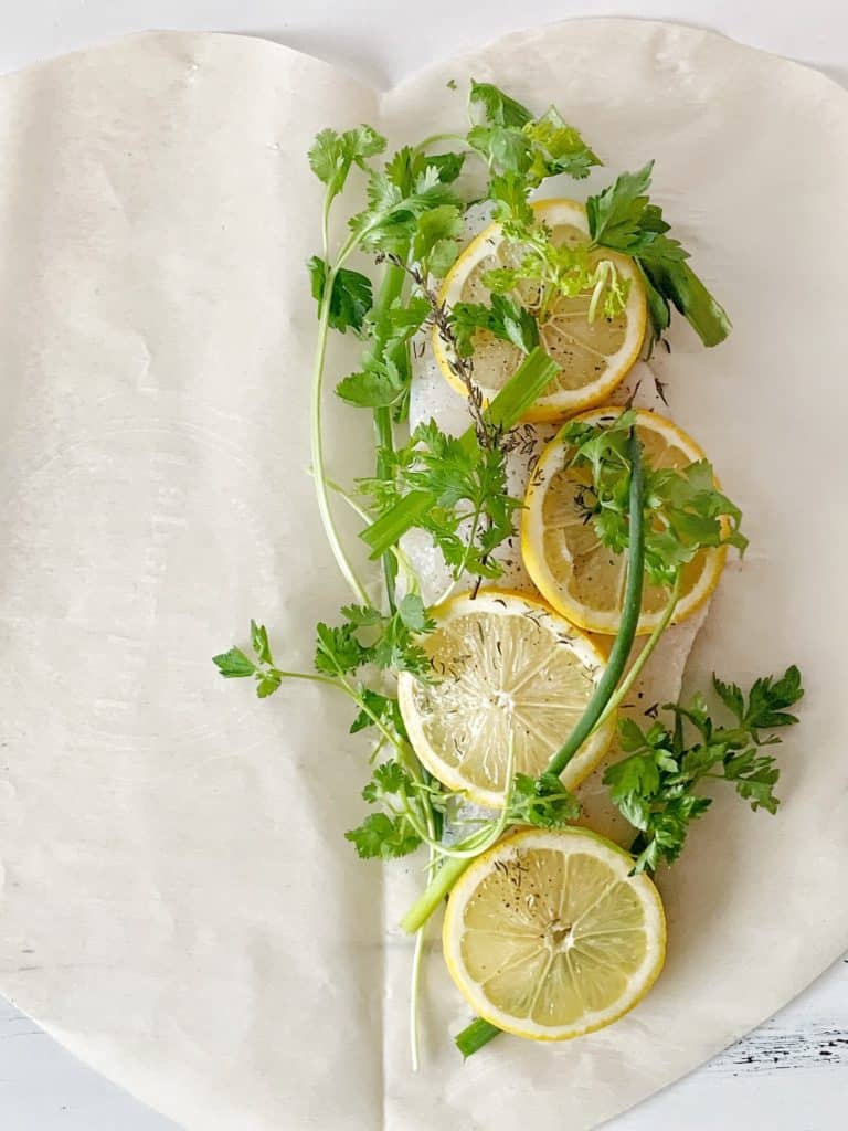 Slice of haddock on a piece of parchment paper with zucchini, lemon, salt and black pepper, cilantro, chives, thyme and parsley