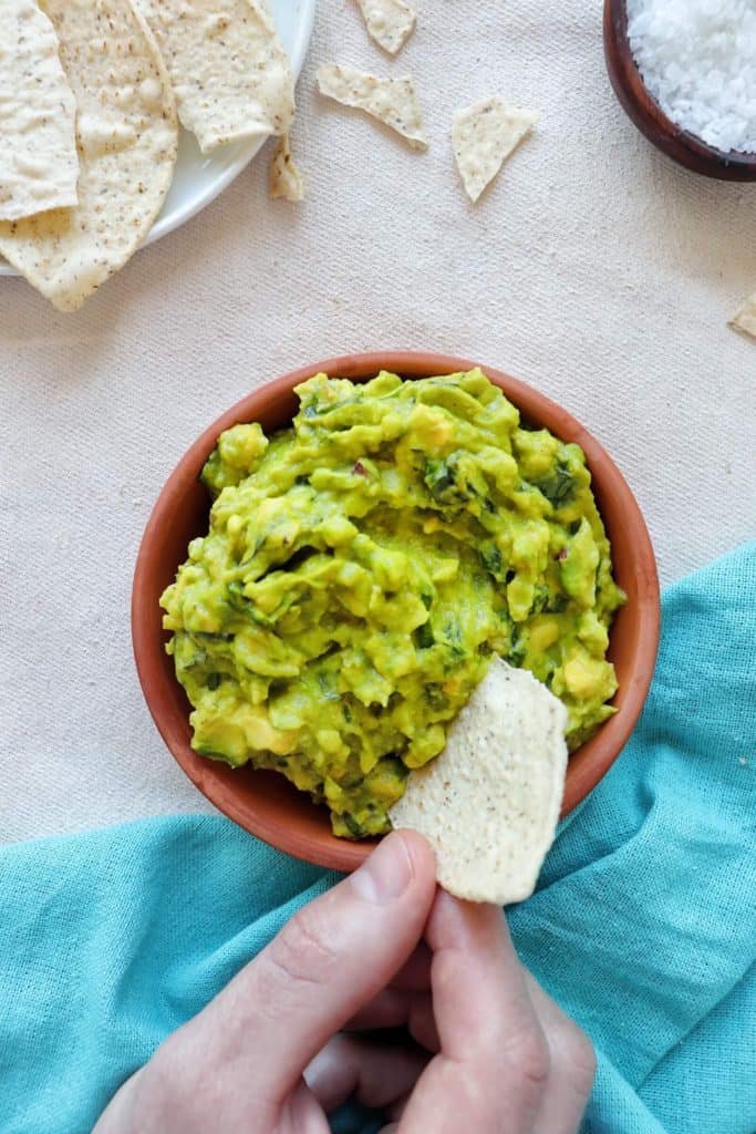 Bowl of Guacamole with a hand dipping a grain-free tortilla chip into the guacamole and blue tea towel
