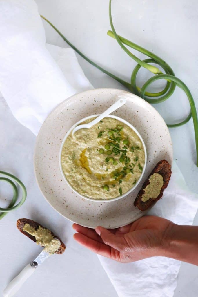 Small plate of Garlic Scape Hummus (AIP) nestled within a larger plate being held by a hand with garlic scapes framing image