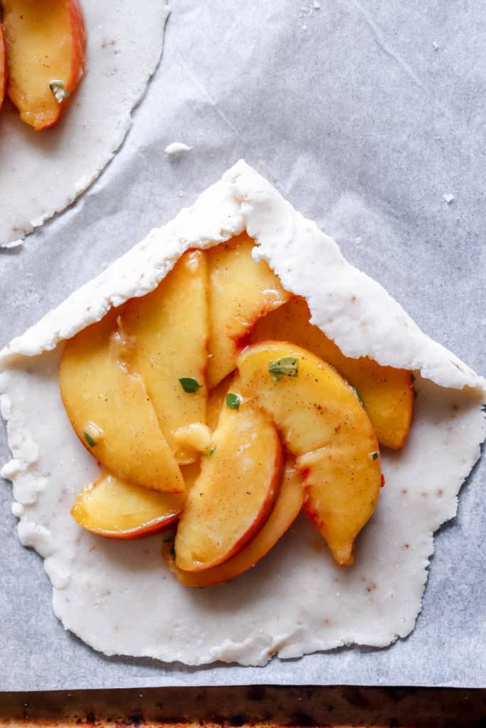 one closeup shot of an unbaked peach galette with the pastry being folded over on a piece of white parchment paper