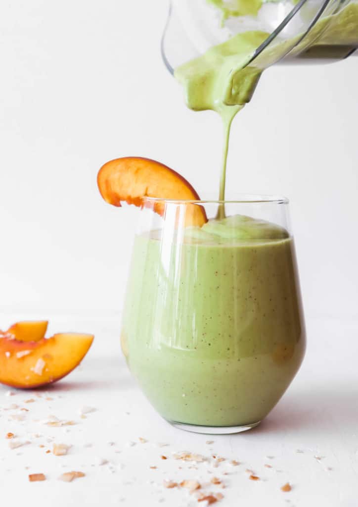 blender pouring peach green smoothie into a glass with a slice of peach on top framed by two slices of peaches on the left a and toasted coconut flakes