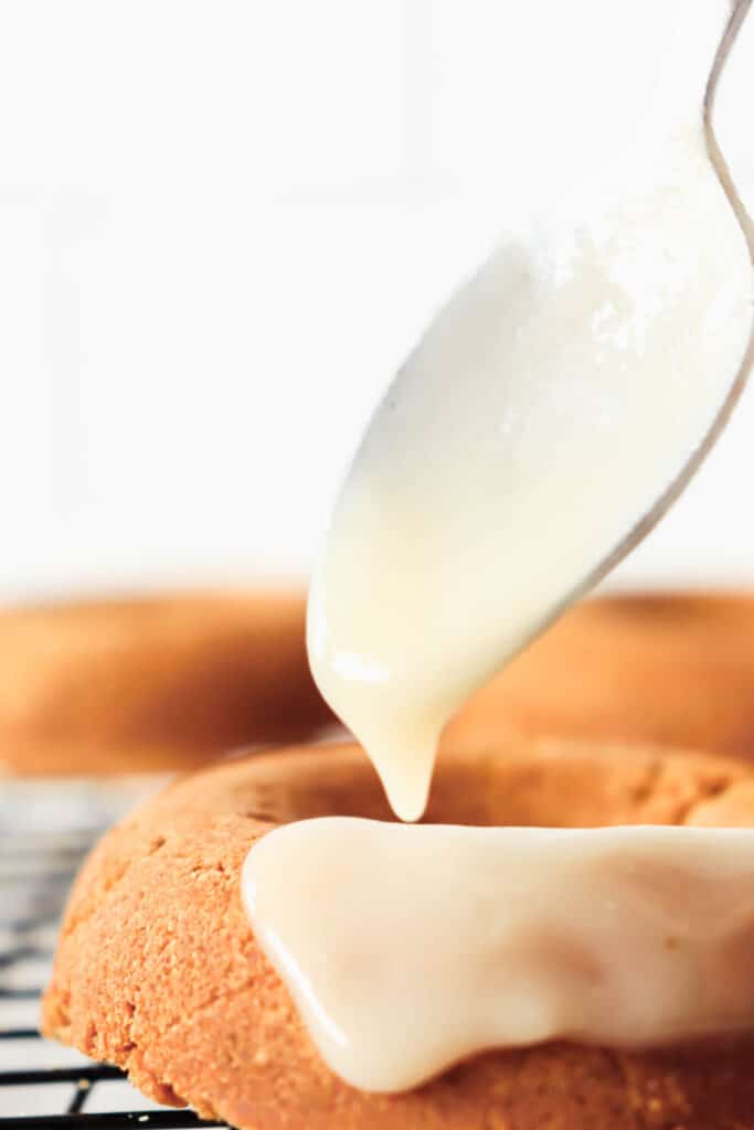 Three pumpkin spice cake donuts on a black cooling rack with a spoon dripping white icing onto one donut