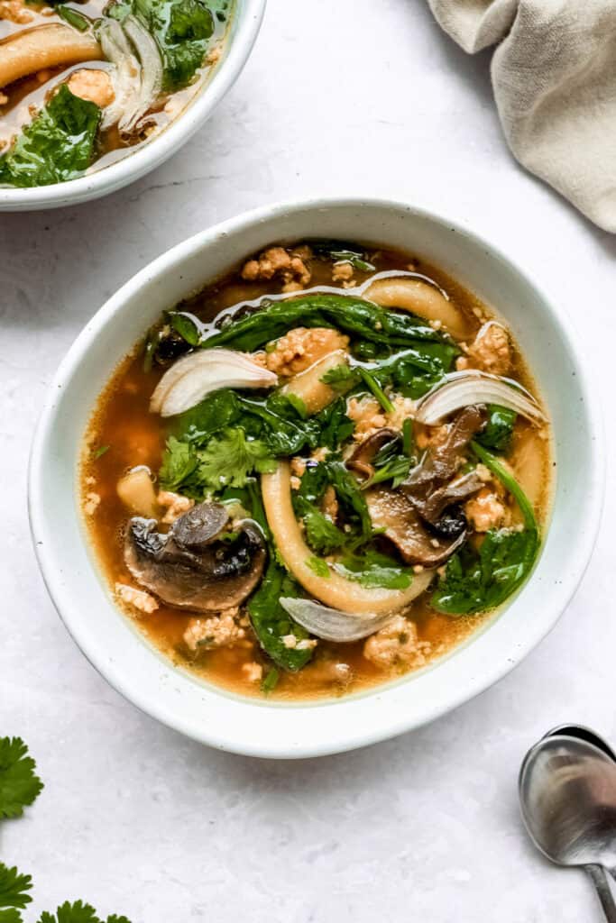 two bowls of ginger-garlic pork noodle soup on a light grey background with two spoons and decorative cilantro