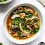 two bowls of ginger-garlic pork noodle soup on a light grey background with two spoons and decorative cilantro