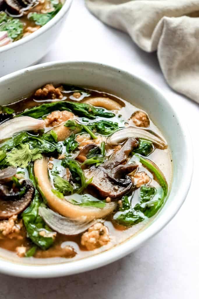 two bowls of ginger-garlic pork noodle soup on a light grey background with two spoons and decorative cilantro