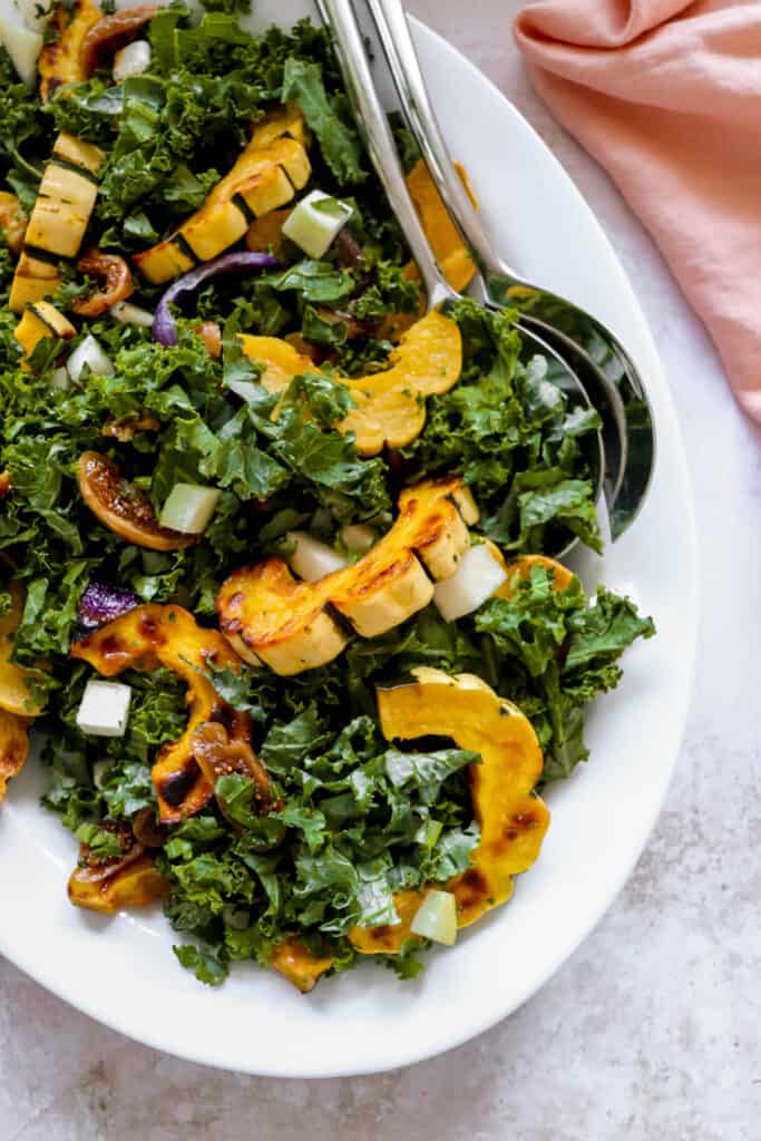 Large white platter with Roasted Delicata Squash and Kale Salad, two serving spoons and a pink cloth napkin