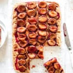 plum sheet cake with a few slices cut on white parchment paper with a white knife and surrounding props