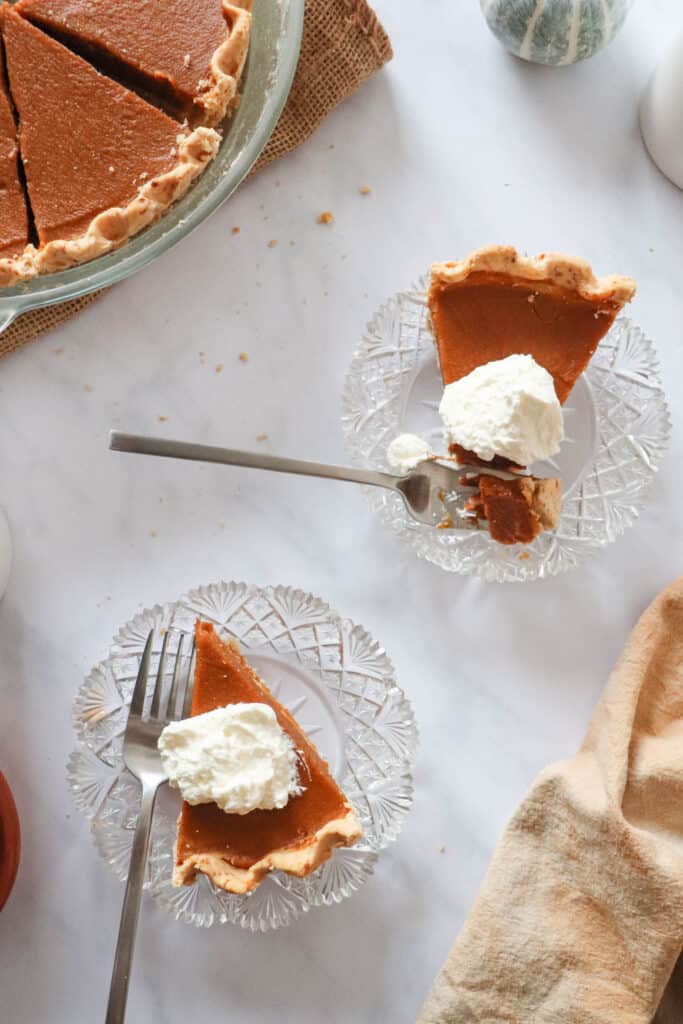 two slices of pumpkin pie with a dollop of coconut whipping cream on each, sitting on crystal plates with two forks, an orange cloth napkin on the bottom right of the image and the remaining slices of pumpkin pie in a glass pie dish on the top left corner