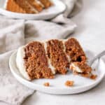 two slices of three layer apple spice cake with white frosting on white plates on top a light brown cloth napkin