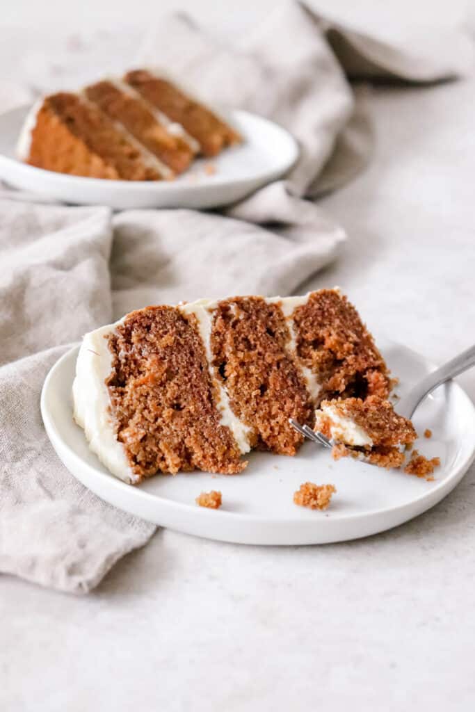 two slices of three layer apple spice cake with white frosting on white plates on top a light brown cloth napkin