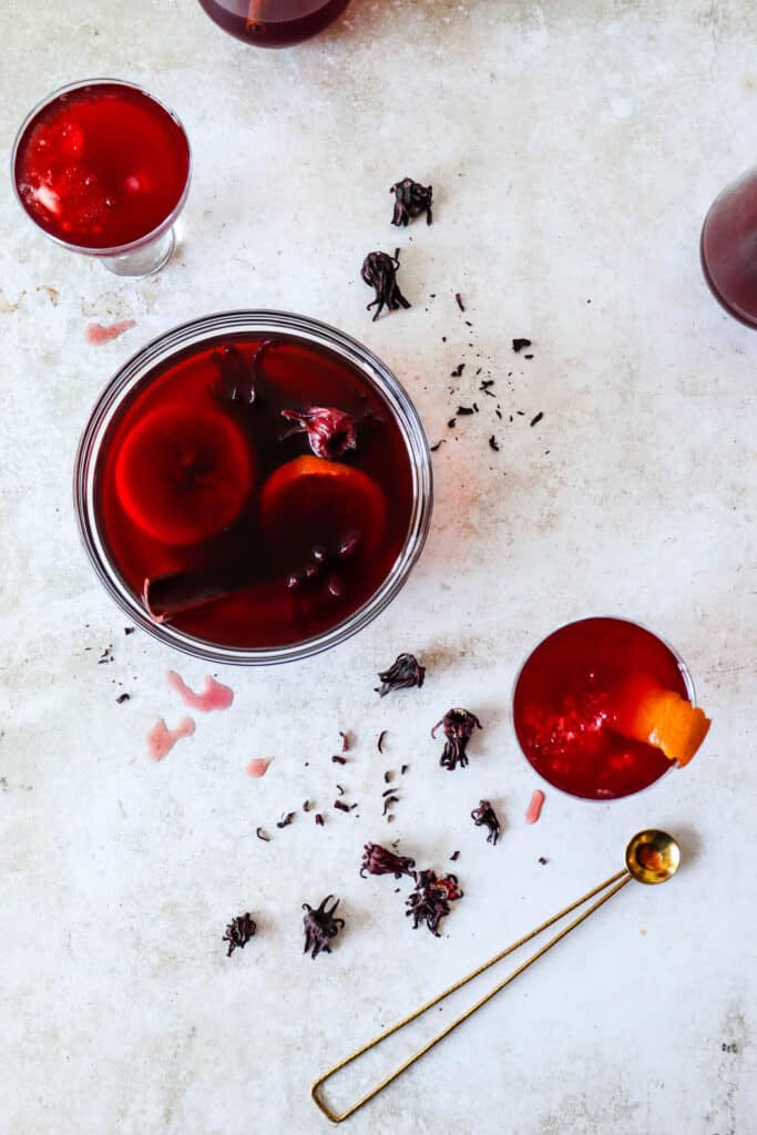 Four glasses with sorrel, with one large glass bowl filled with sorrel, cinnamon sticks and sliced oranges, scattered dried hibiscus flowers and brass cocktail spoon