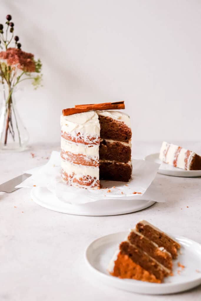 three layer cake with white frosting and cinnamon sticks on top with two small white plates with a slice of cake on each