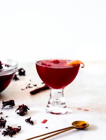 Glass of sorrel garnished with an orange peel next to a glass bowl of sorrel and scattered dried hibiscus flowers and brass cocktail spoon