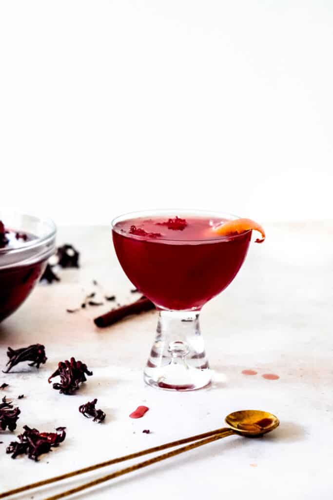 Glass of sorrel garnished with an orange peel next to a glass bowl of sorrel and scattered dried hibiscus flowers and brass cocktail spoon