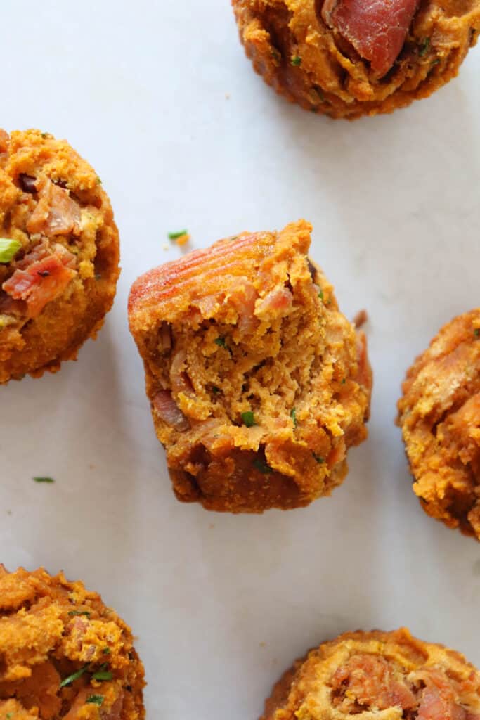 five sweet potato, bacon and chive muffins on a white background with a bite taken out of one