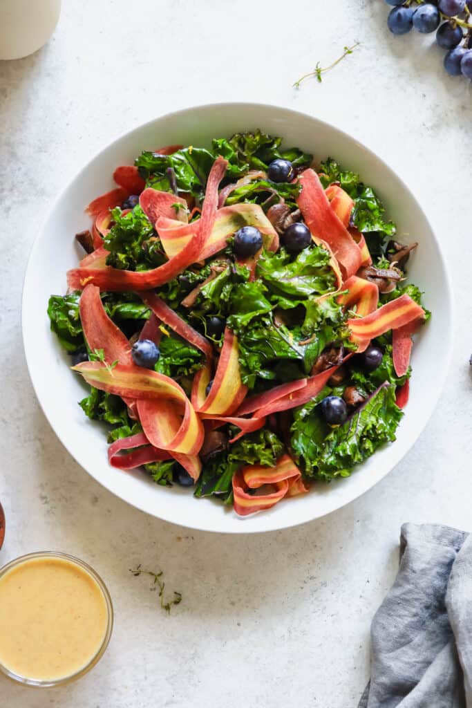 White bowl with sauteed kale, ribbons of carrots, coronation grapes, mushrooms and red onions on a grey speckled background with a small bowl of yellow salad dressing and a grey cloth napkin 