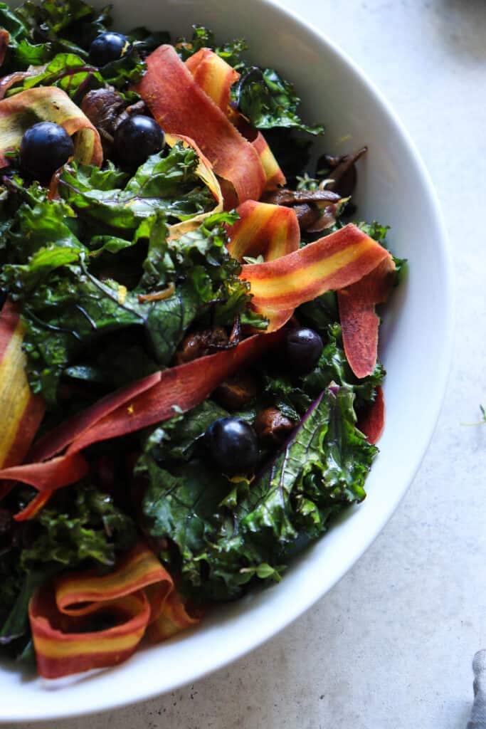 White bowl with sauteed kale, ribbons of carrots, coronation grapes, mushrooms and red onions on a grey speckled background