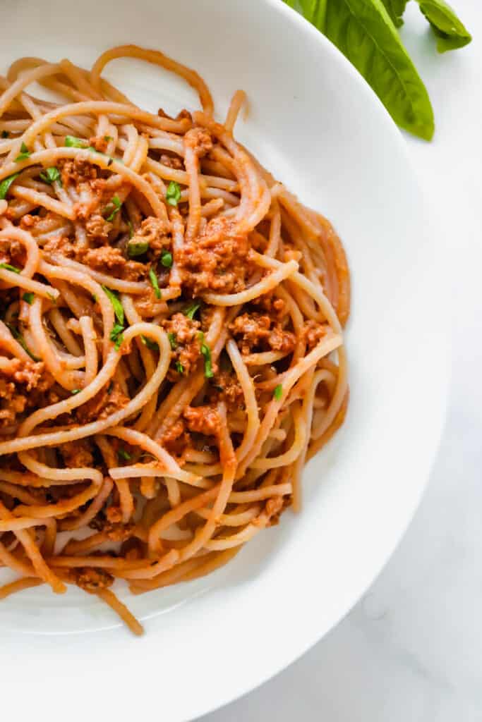 White bowl with cassava flour spaghetti and meat sauce topped with chopped basil against a white backdrop with a bunch of fresh basil in the top right corner
