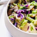 Cream coloured bowl with Sautéed Veggies (purple cabbage, yellow carrots, broccoli and cauliflower) on a white background with a beige cloth napkin