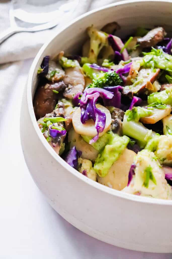 Cream coloured bowl with Sautéed Veggies (purple cabbage, yellow carrots, broccoli and cauliflower) on a white background with a beige cloth napkin