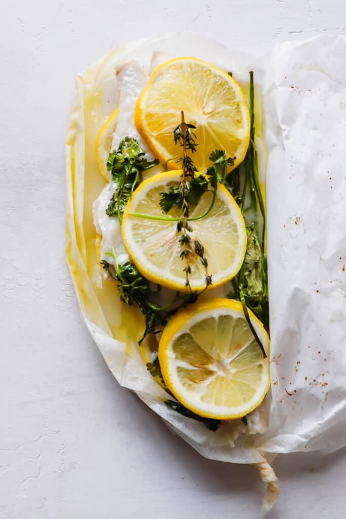 haddock fillet topped with sliced lemon, thyme and cilantro in a packet of parchment paper on a white backdrop 
