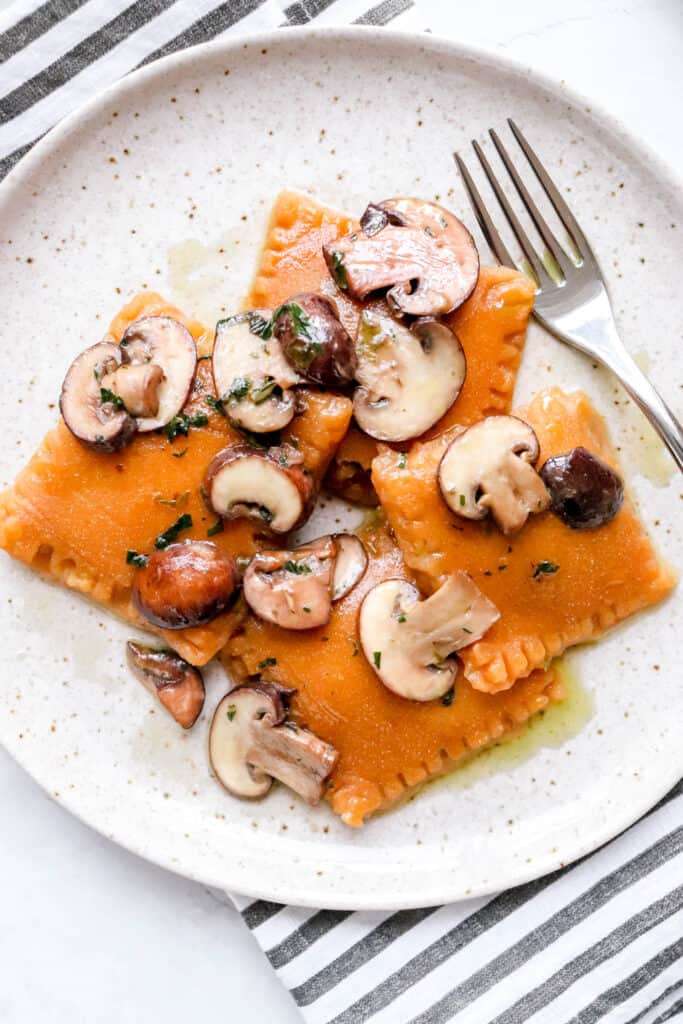 speckled plate with three sweet potato raviolis, topped with sliced mushrooms and spinach and a fork lying on top a grey and white striped cloth napkin