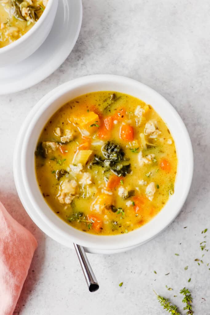 two white bowls of soup on white saucers with carrots, chopped kale, ground turkey and fresh thyme with a spoon next to one bowl and a pink cloth napkin