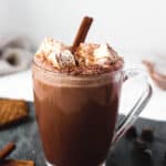 glass mug of hot chocolate topped with marshmallows on a grey slate board, with garnishing of cookies and cinnamon sticks