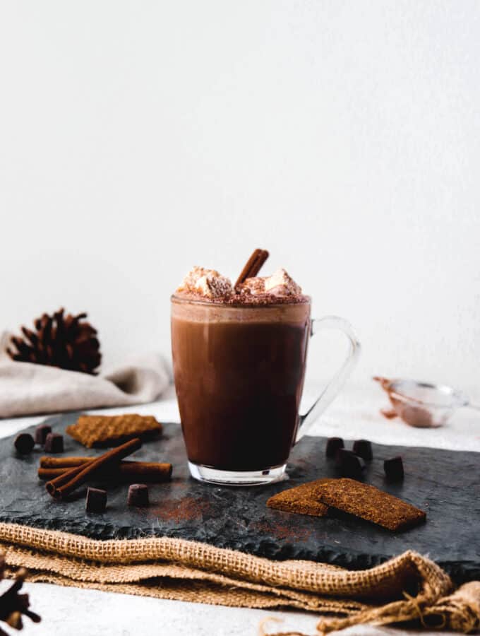glass mug of hot chocolate topped with marshmallows on a grey slate board on top a burlap mat, with garnishing of cookies and cinnamon sticks and acorns