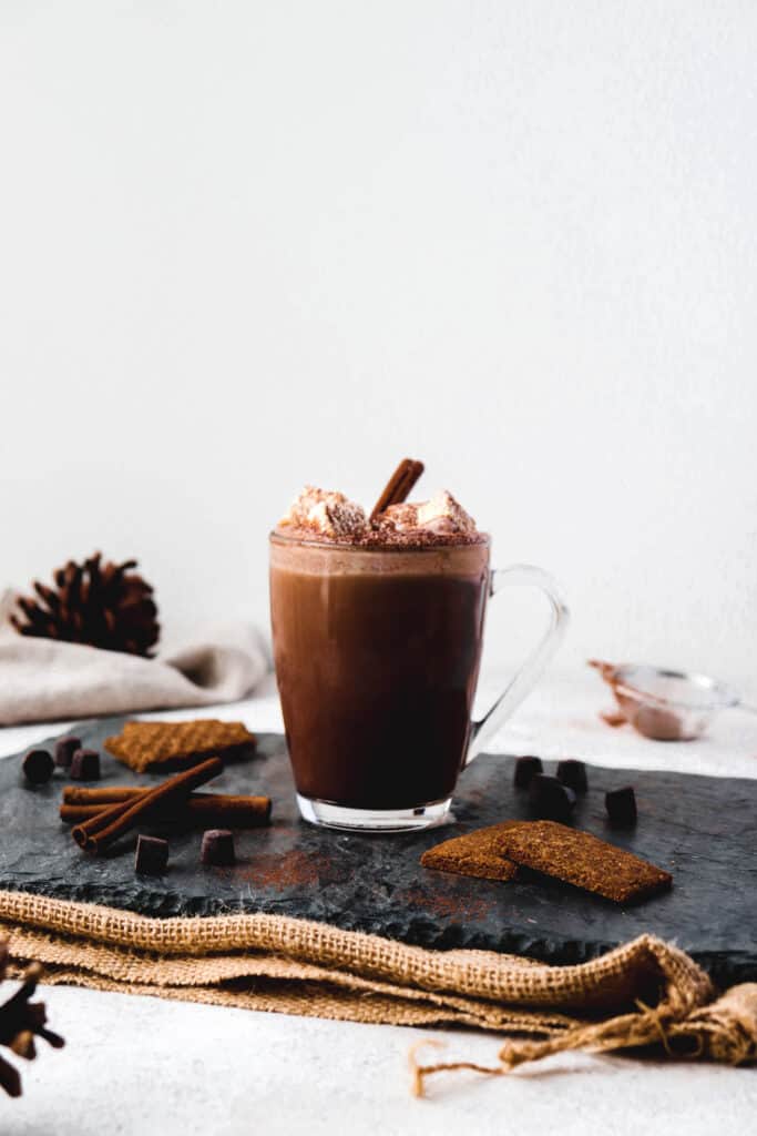 glass mug of hot chocolate topped with marshmallows on a grey slate board on top a burlap mat, with garnishing of cookies and cinnamon sticks and acorns