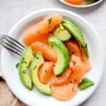 two white plates with segments of grapefruit and avocado with sea salt, fresh chopped basil and a fork and an orange cloth napkin