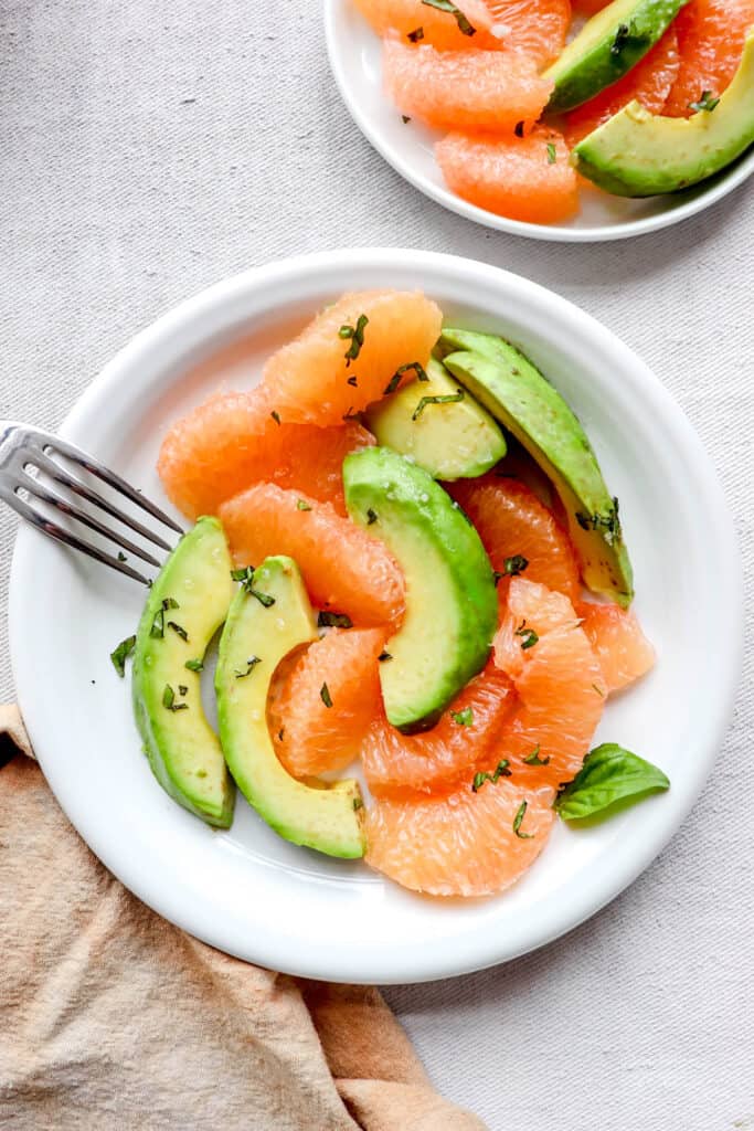 two white plates with segments of grapefruit and avocado with sea salt, fresh chopped basil and a fork and an orange cloth napkin