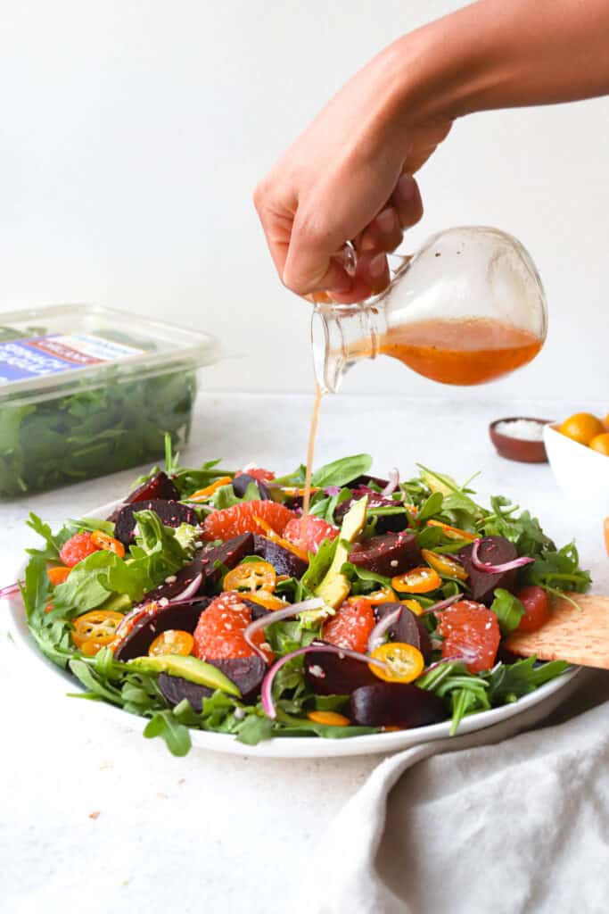 hand pouring vinaigrette over white platter with arugula, spinach, grapefruits, sliced beets and avocado with plastic package of greens in the background