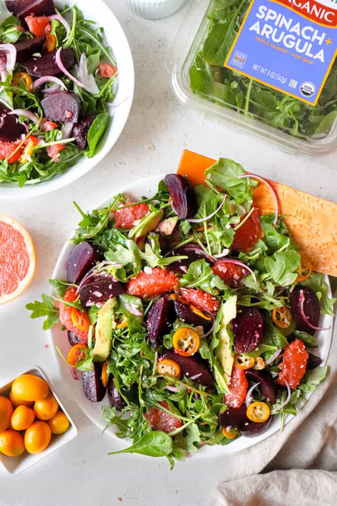 two white platters with arugula, spinach, grapefruits, sliced beets and avocado, with wooden salad forks, a white bowl with salad dressing, a small bowl of salt, a bowl of kumquats and a package of spinach and arugula