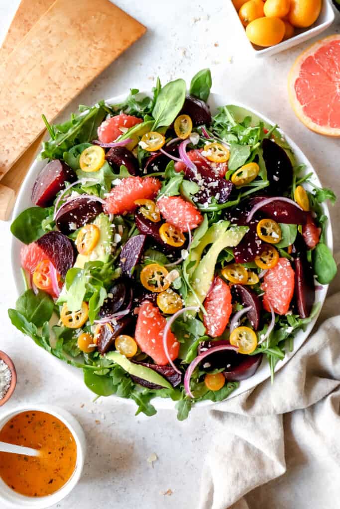 white platter with arugula, spinach, grapefruits, sliced beets and avocado, with wooden salad forks, a white bowl with salad dressing, a small bowl of salt, a bowl of kumquats, one half of a grapefruit and a package of spinach and arugula