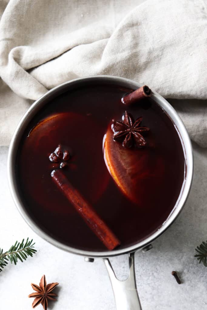 saucepan with pomegranate juice, orange slices, cinnamon sticks, cloves and star anise against a white background with beige napkins and garnish of pine leaves and star anise