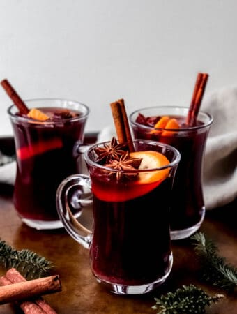 three glass mugs of mulled wine mocktail with garnishes of orange slices, cinnamon sticks and star anise on a dark brown serving tray with pine leaves and cinnamon sticks