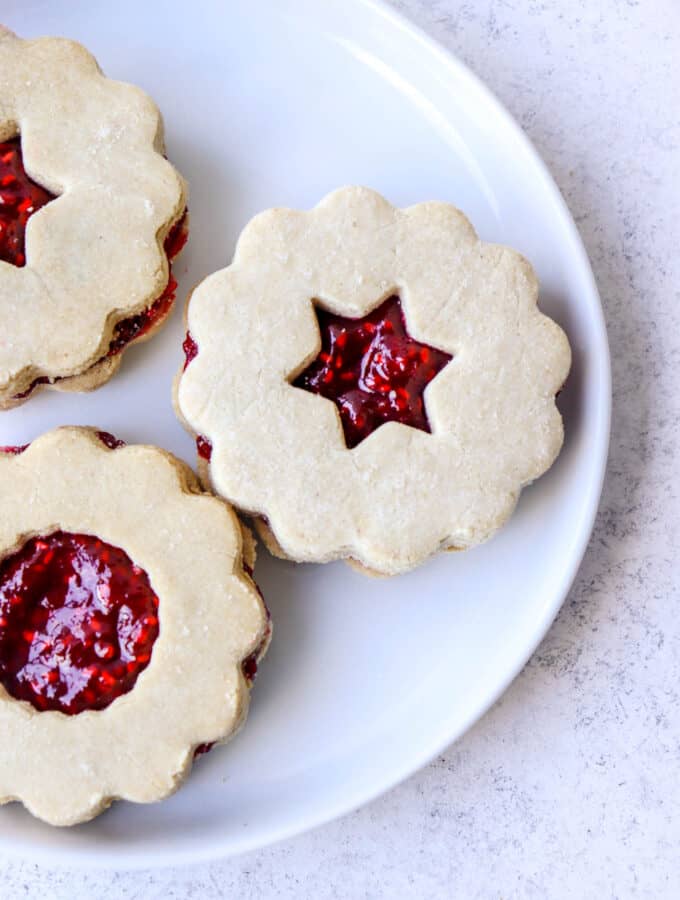 three raspberry filled linzer cookies on a white plate on a grey backdrop