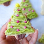 hand holding christmas tree cookie decorated with green, brown, pink and yellow icing with more cookies in the background, blurred