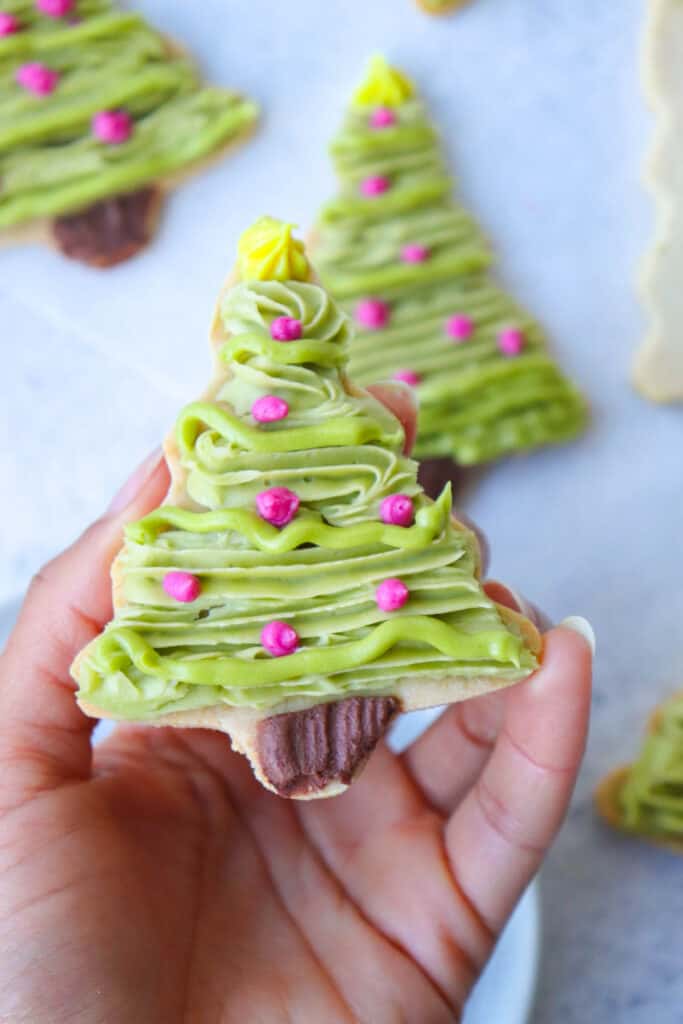hand holding christmas tree cookie decorated with green, brown, pink and yellow icing with more cookies in the background, blurred