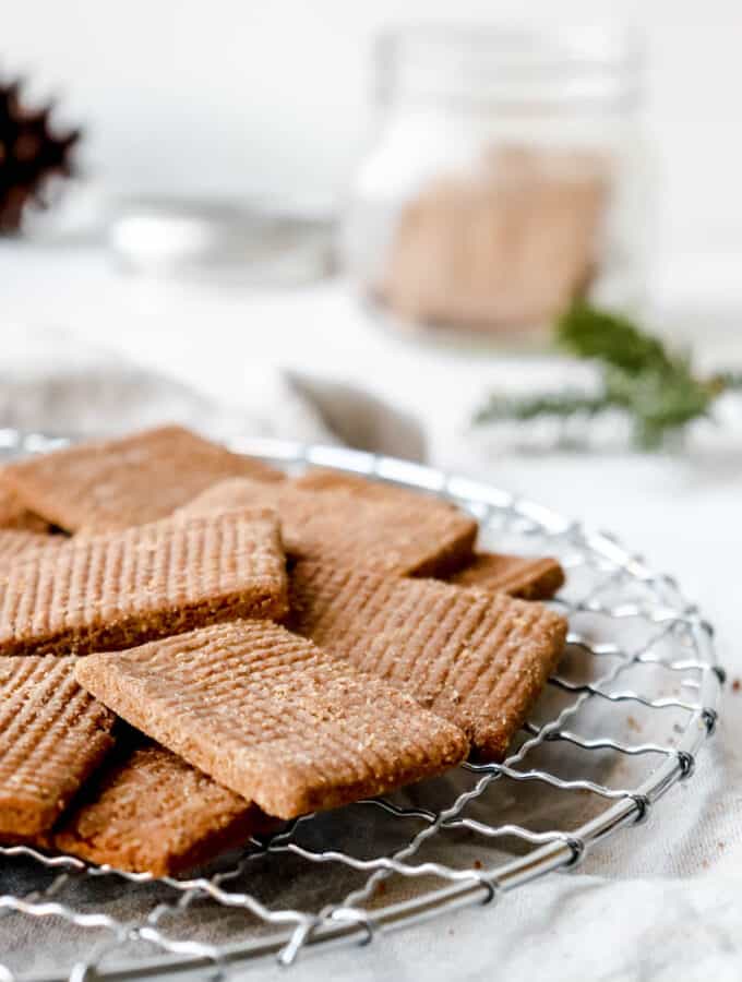 speculaas on a metal round cooling rack against a grey backdrop with a beige cloth napkin and a jar of cookies blurred in the background