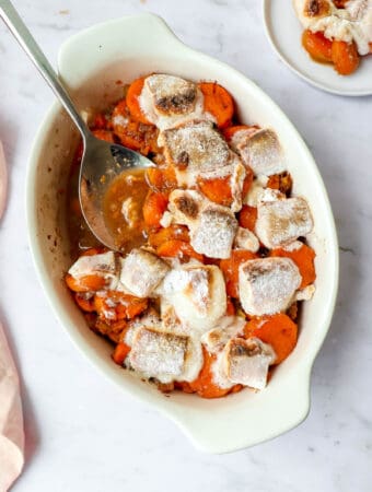cream colored oval casserole dish with sweet potatoes, cinnamon, crushed pineapple and roasted marshmallows with a serving spoon and a side plate of sweet potato casserole
