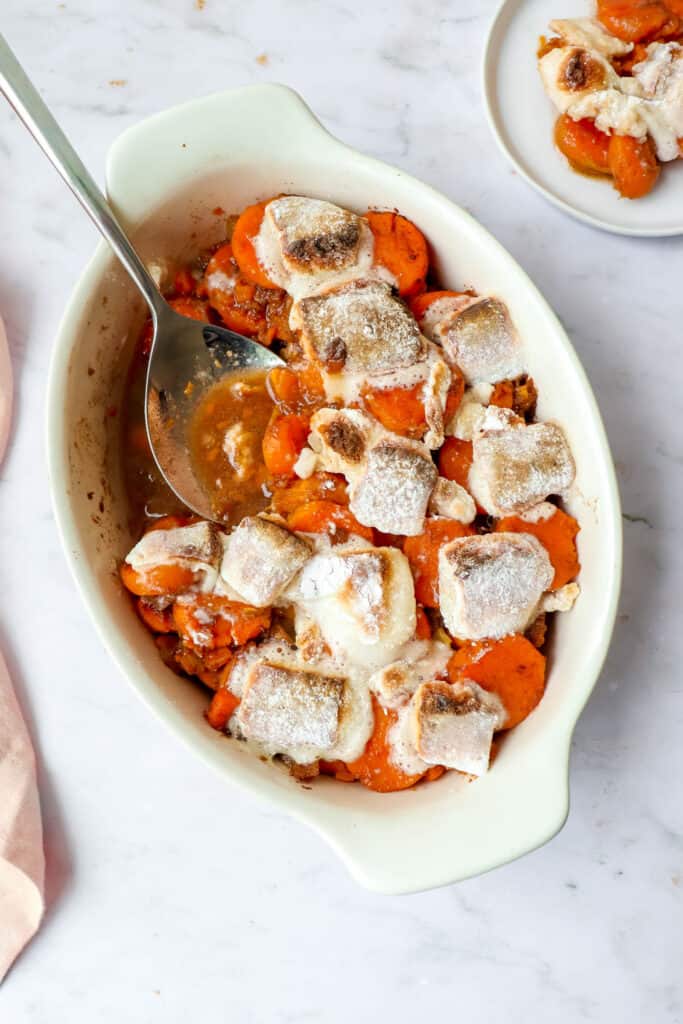 cream colored oval casserole dish with sweet potatoes, cinnamon, crushed pineapple and roasted marshmallows with a serving spoon and a side plate of sweet potato casserole