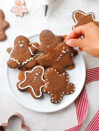 hand reaching into white plate with gingerbread people cookies iced with white icing on a grey background with a red and white ribbon, a gingerbread man cookie cutter, scattered gingerbread cookies, an angel ornament and an icing tip