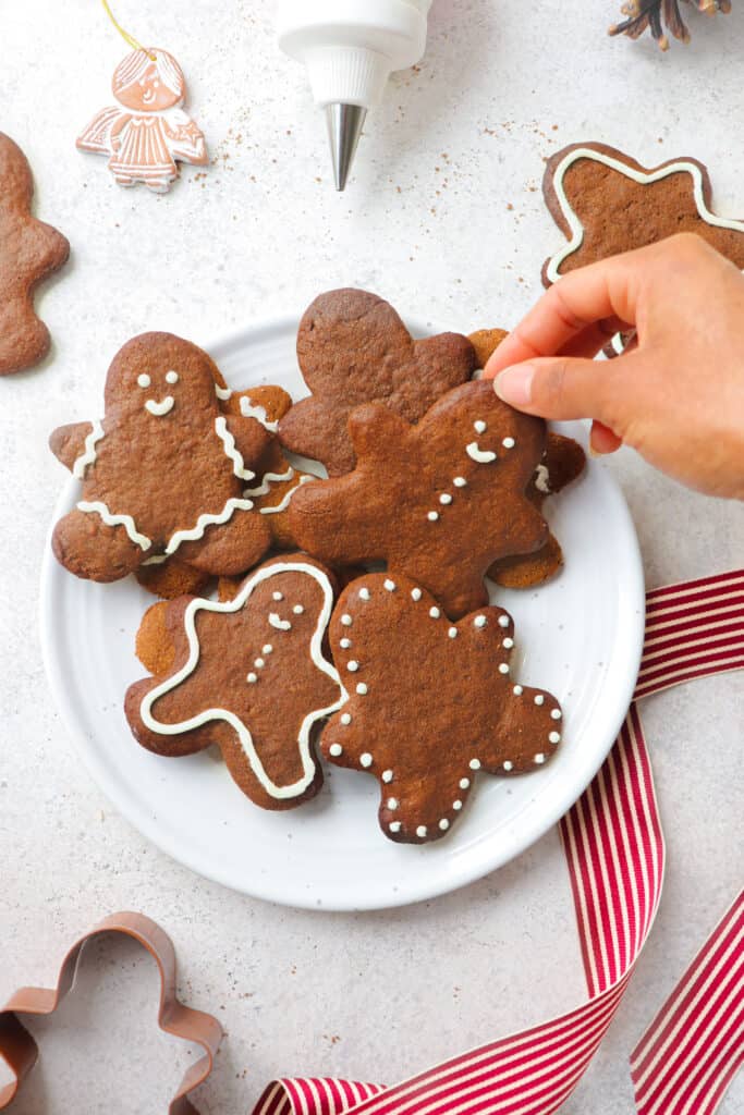 hand reaching into white plate with gingerbread people cookies iced with white icing on a grey background with a red and white ribbon, a gingerbread man cookie cutter, scattered gingerbread cookies, an angel ornament and an icing tip