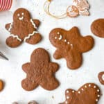 gingerbread people cookies iced with white icing on a grey background with a red and white ribbon, a gingerbread man cookie cutter, scattered gingerbread cookies, an angel ornament and an icing tip