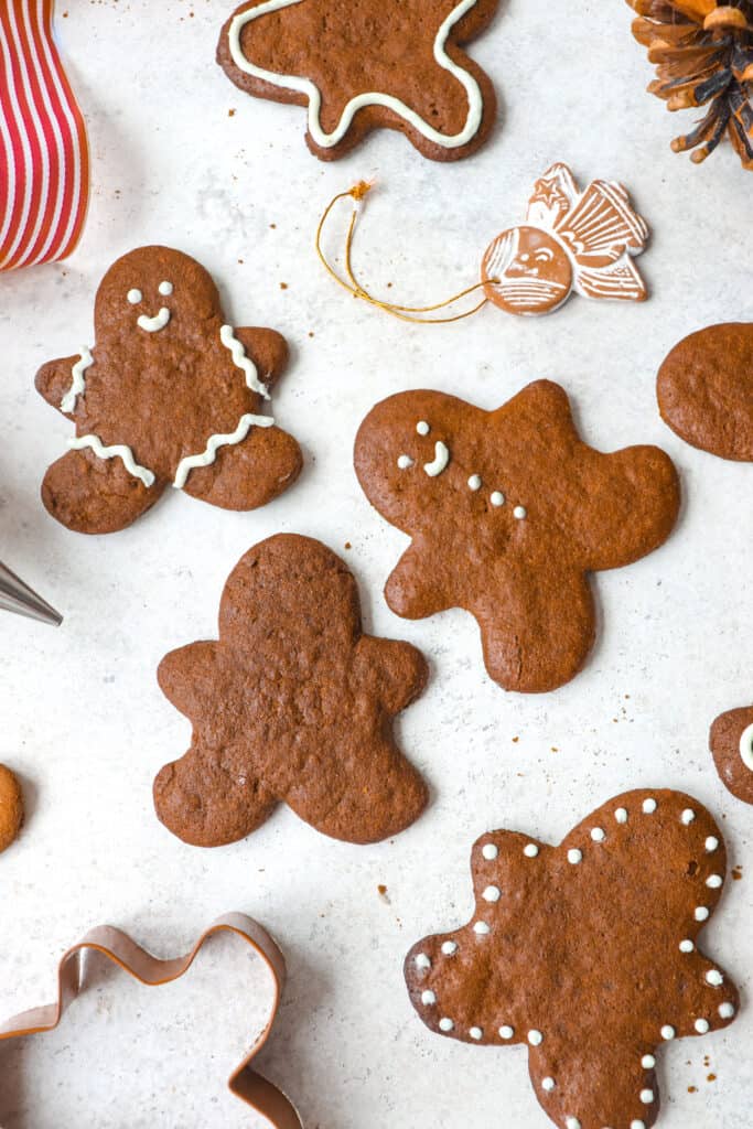 gingerbread people cookies iced with white icing on a grey background with a red and white ribbon, a gingerbread man cookie cutter, scattered gingerbread cookies, an angel ornament and an icing tip